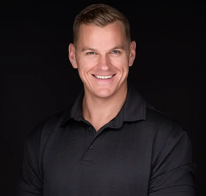 Smiling man with short light hair wearing a black collared shirt stands against a plain black background, capturing the polished look often seen in professional headshots in Raleigh Nc