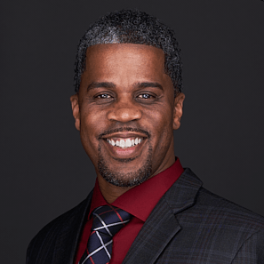 A man with short curly hair and a beard smiles in his professional headshot, wearing a dark suit, red shirt, and checkered tie against a dark background.