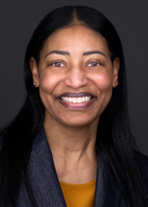 An African American lady smiles for a professional headshot while wearing a blue jackets with sparkles and a mustard colored top at the portrait studio of Sarah Anne Wilson in Cary NC