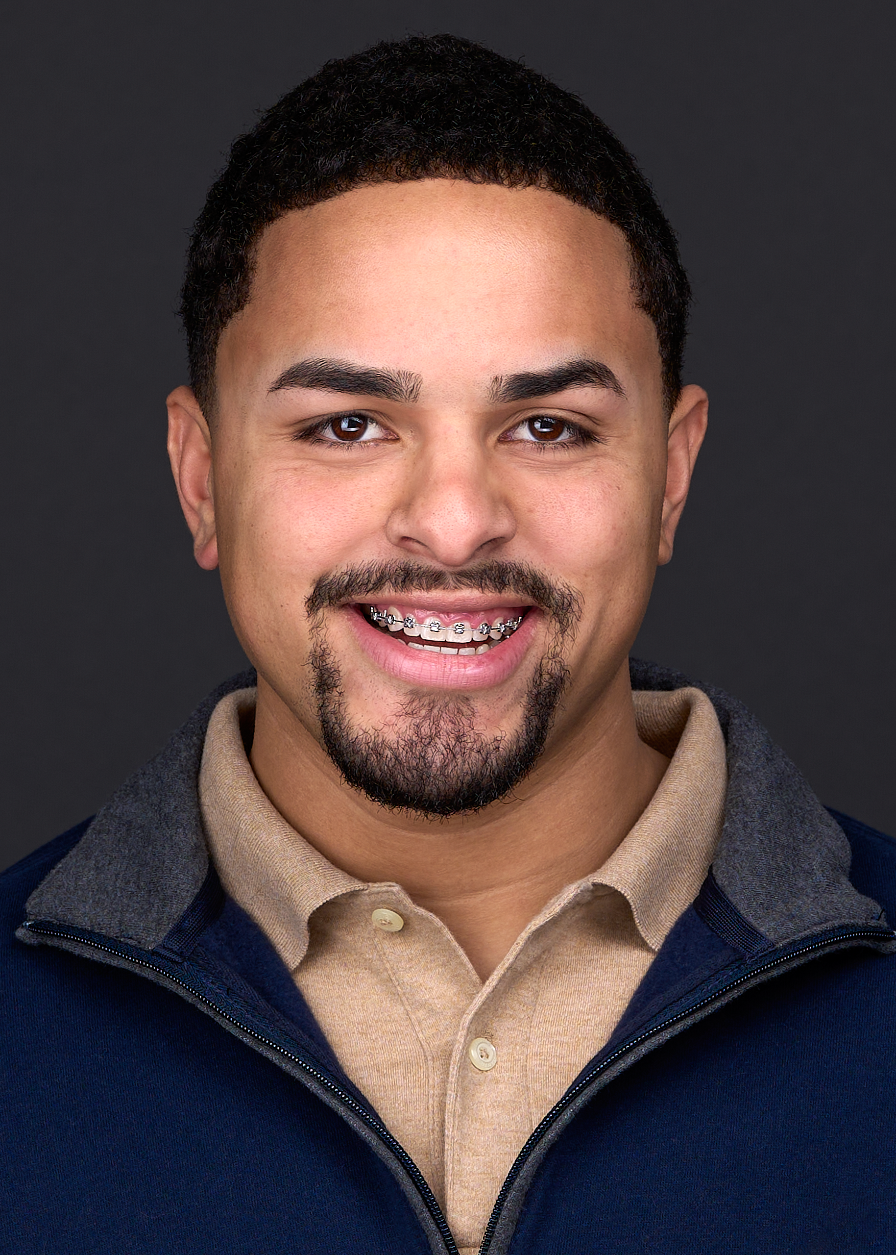 A young man with a beard and mustache poses for a professional headshot while wearing a beige shirt and blue jacket in Cary NC