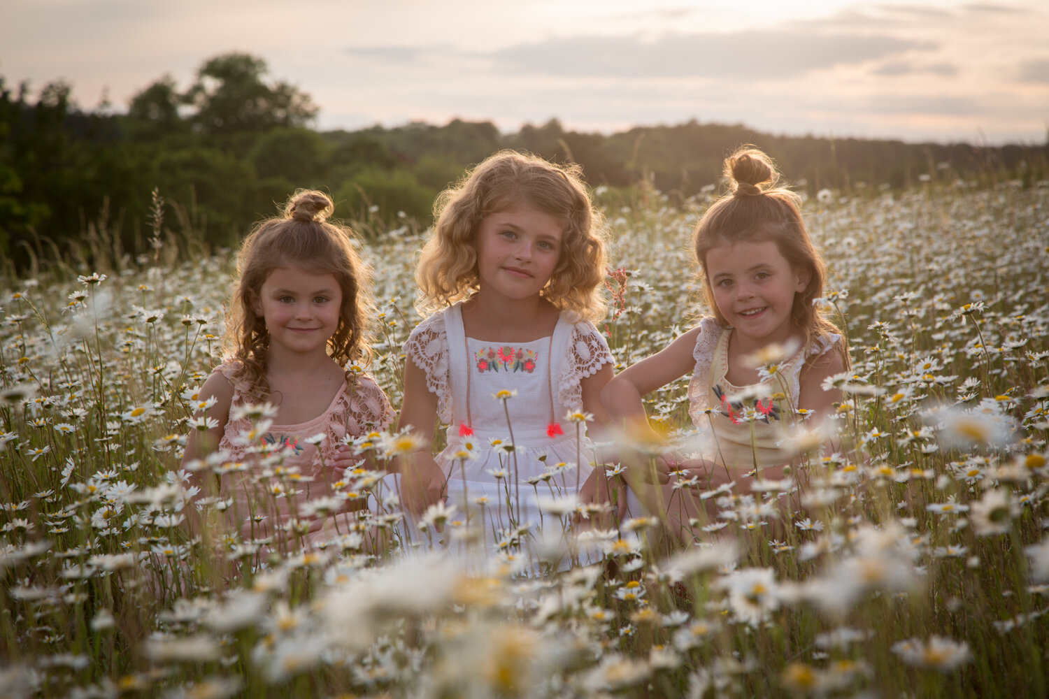 Three young girls sit among blooming white daisies in a field at sunset, capturing the magic of the golden hour with greenery in the background.