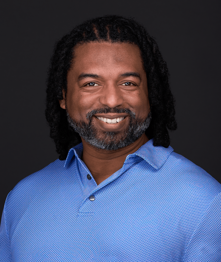 A person with shoulder-length braided hair, a beard with gray streaks, and a big smile, wearing a blue collared shirt, is looking at the camera against a black background—perfect for professional business headshots.