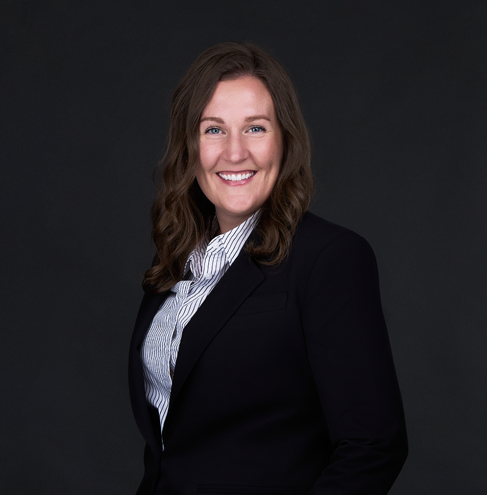 A woman with long brown hair wearing a navy blazer and striped shirt smiles in front of a plain dark background, capturing the essence of professional women's headshots.