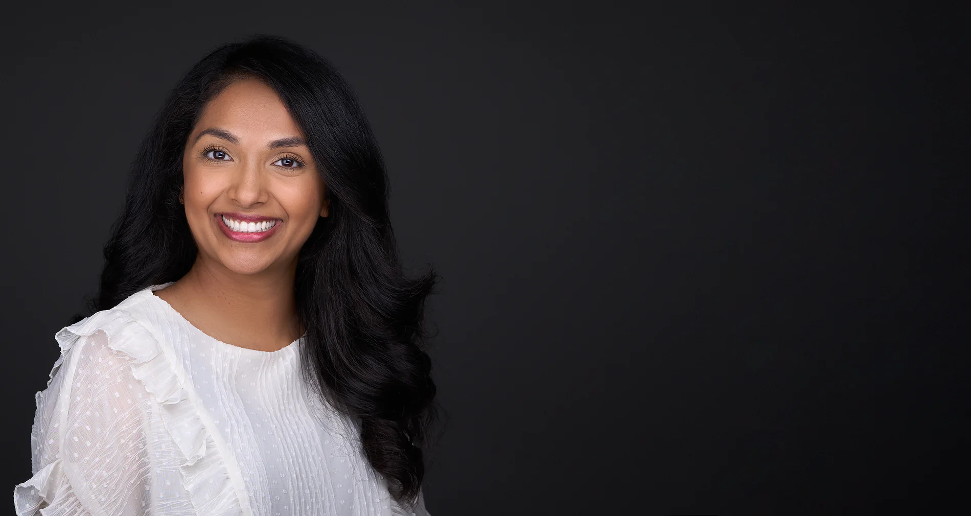 A woman with long black hair and a white blouse smiles confidently in her women's headshot against a dark background.