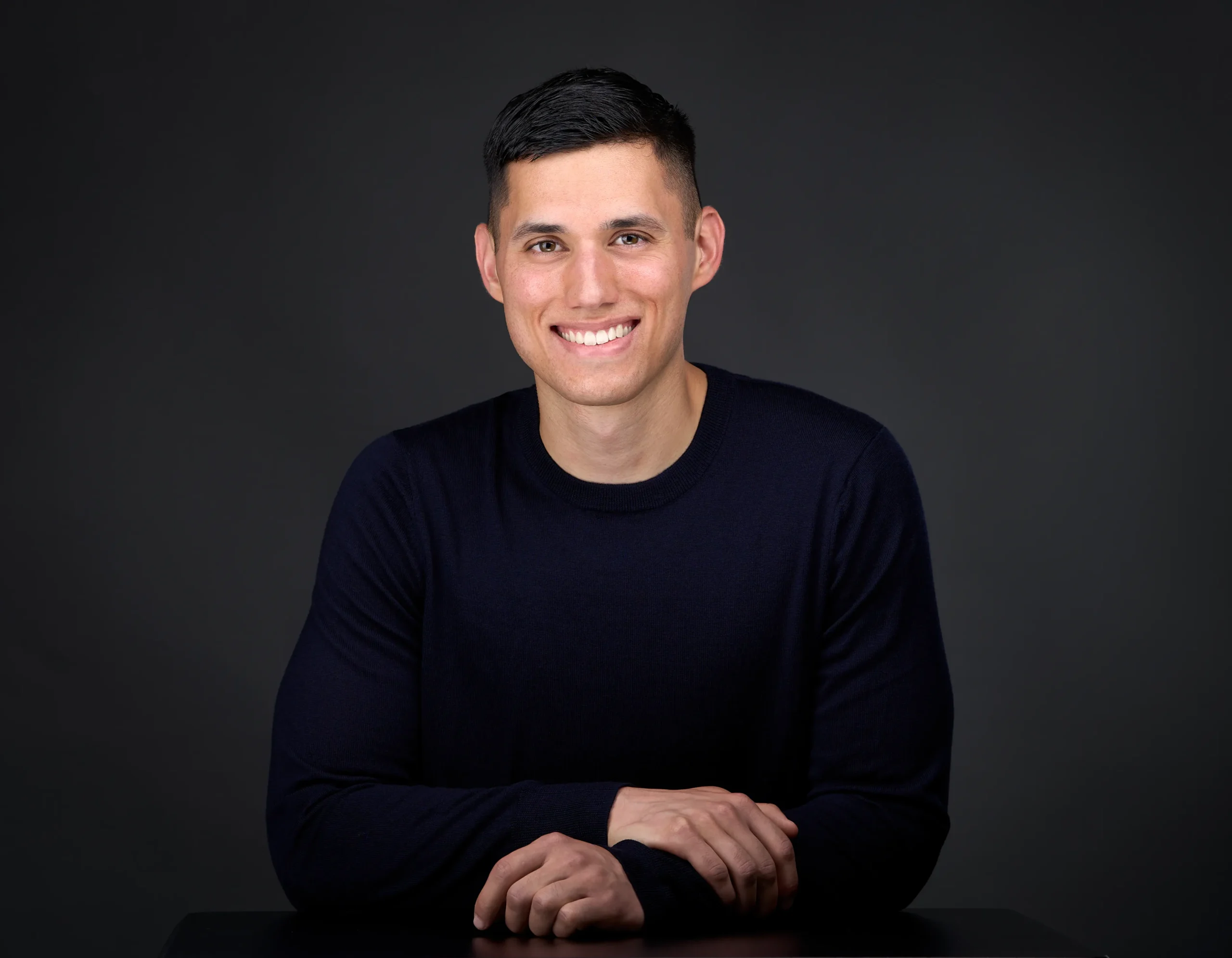 A young man poses while leaning on a table for a dating profile image. He's wearing navy crew neck sweater