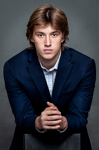 A person with medium-length hair is wearing a dark blazer over a light checkered shirt and clasping their hands while looking directly at the camera against a dark background, perfectly capturing the essence of branding photography.