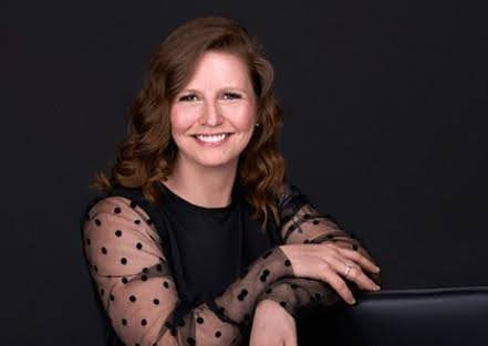 A woman with wavy hair, wearing a black top with sheer, polka-dotted sleeves, smiles while leaning on a black surface against a dark background. The image exudes professionalism, making it perfect for branding photography.