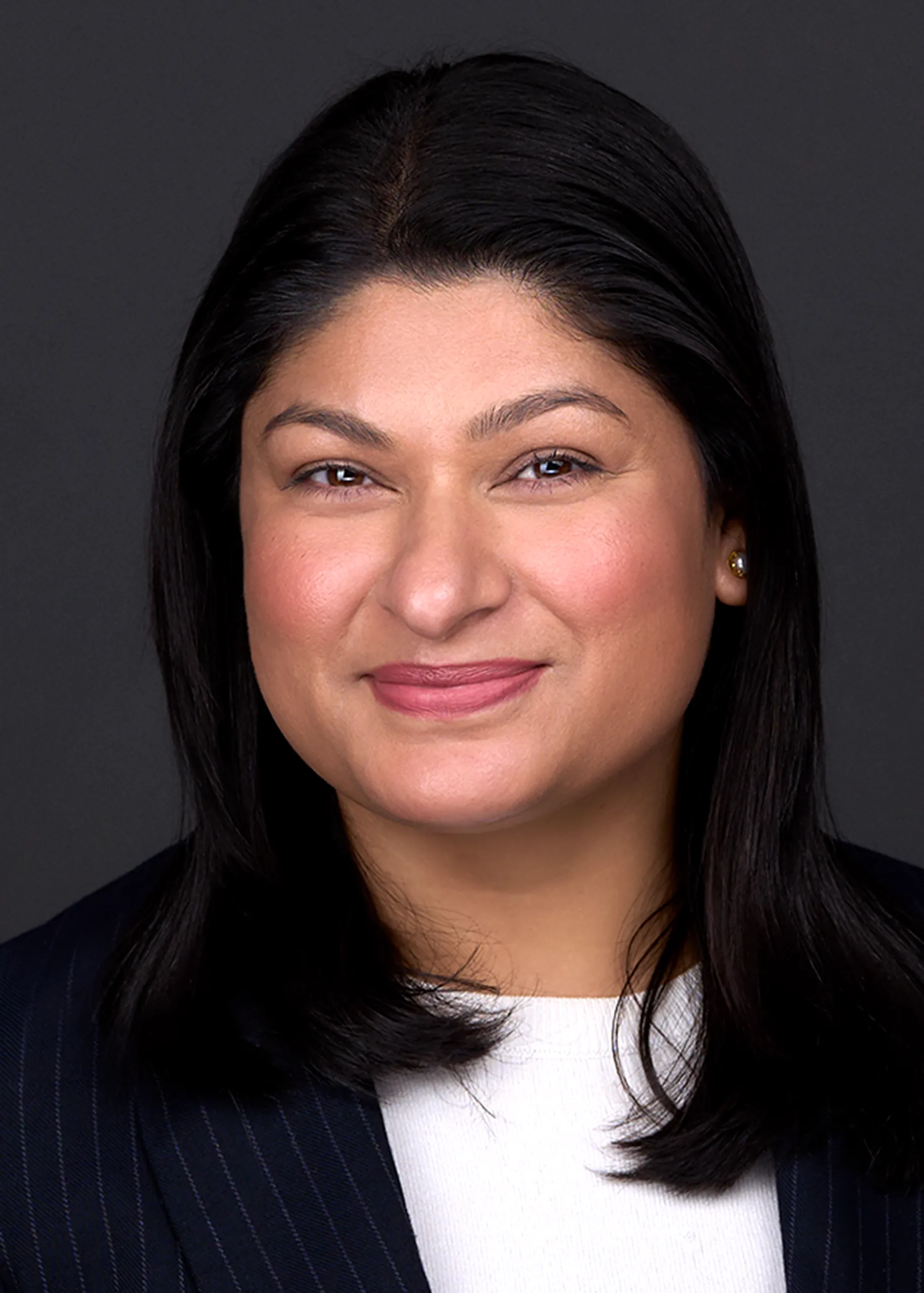 A woman with dark hair wearing a dark blazer and white shirt smiles at the camera against a black background, making for a perfect professional headshot.