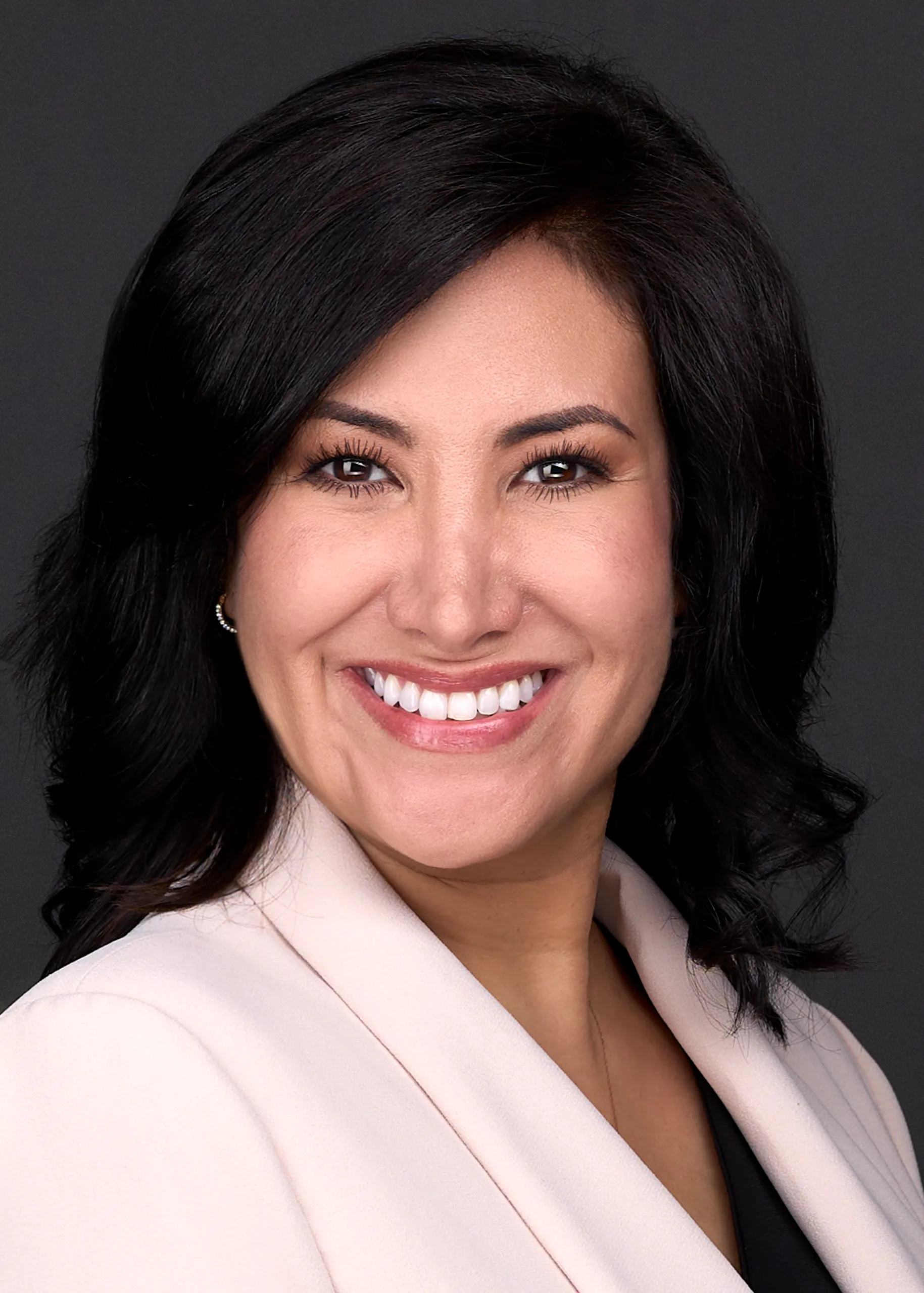 A person with shoulder-length black hair and a light-colored blazer smiles in front of a dark background, perfect for professional headshots.