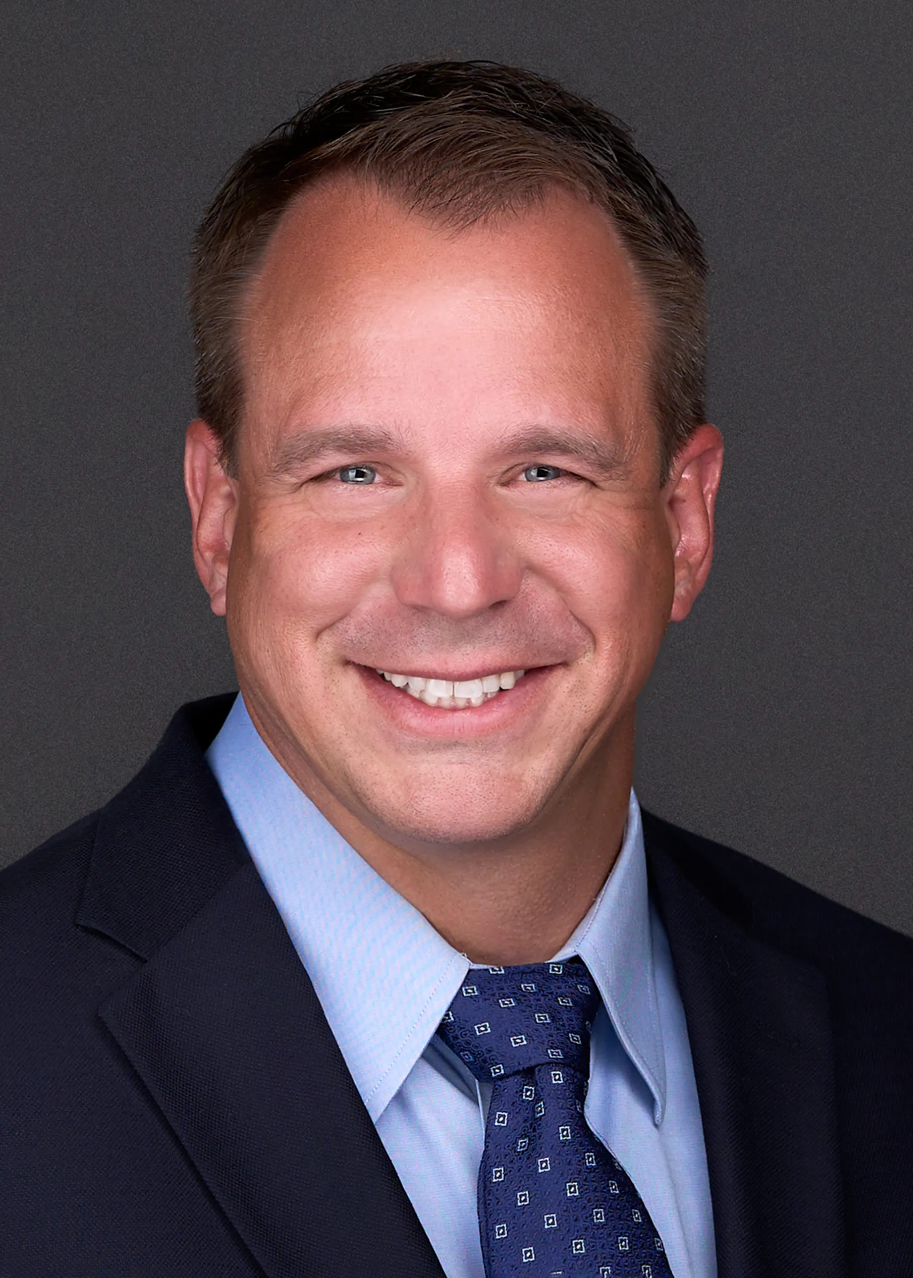 A professional business man dressed in a navy suite and light blue shirt smiles in front of the camera while sitting for a professional headshot in Raleigh NC