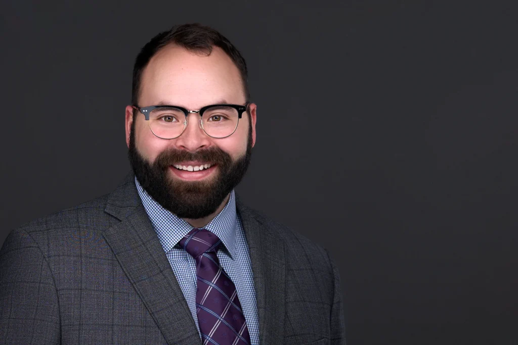 A business man with glasses, a beard, and short hair, wearing a gray suit, checkered shirt, and purple tie stands smiling against a dark background—perfect for professional headshots.