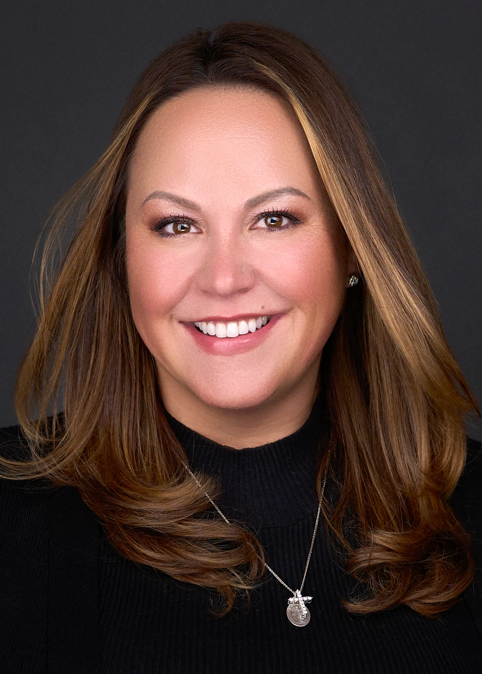 A young woman with long brown hair wearing a black sweater smiles warmly as she poses for a professional headshot for her business in Raleigh Nc