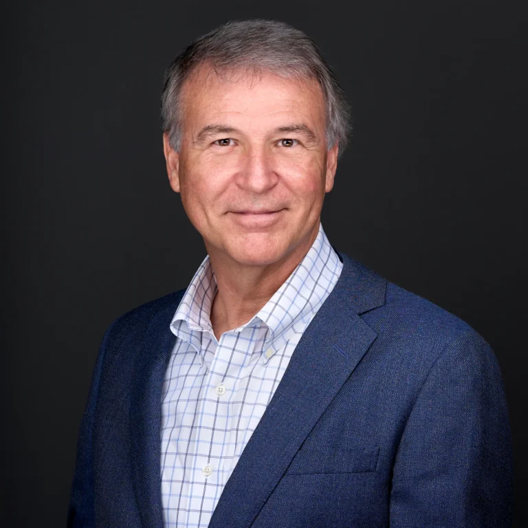 A man with gray hair wearing a blue jacket and plaid white shirts stands in front of a balck backdrop while smiling for his professional headshots in Raleigh Nc