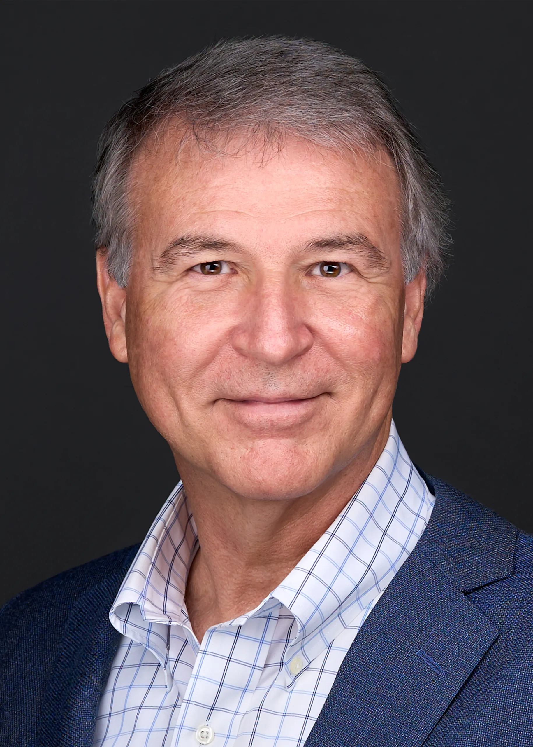 A man with gray hair wearing a blue jacket and plaid white shirts stands in front of a balck backdrop while smiling for his professional headshots in Raleigh Nc