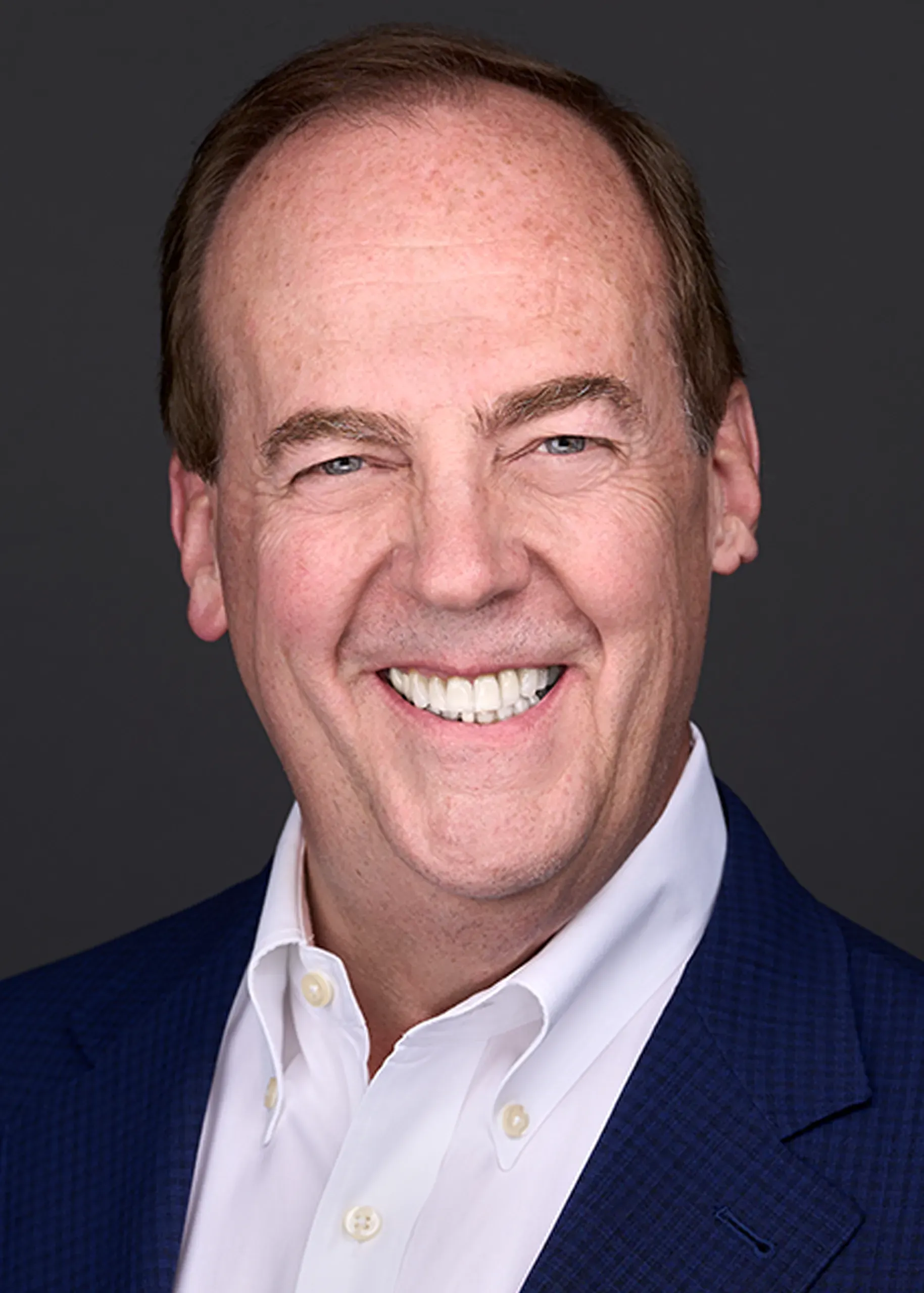 A man dresed in a blue suit and white shirt casually smiles as he stands in front of a dark gray backdrop while being photographed for his professional headshot