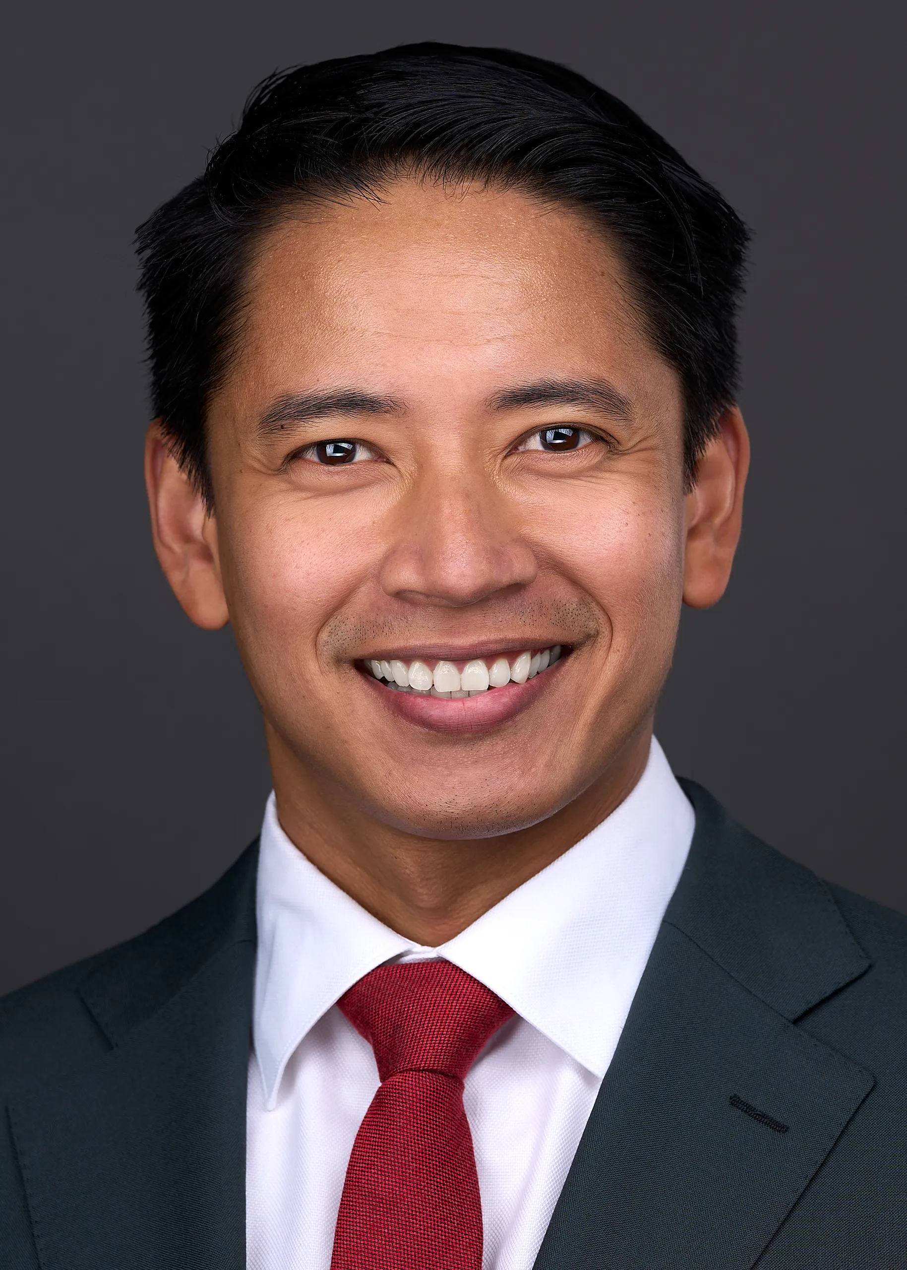 Young man wearing a red tie and gray jacket stands in front of a gray backdrop while posing for a professional headshot in Cary Nc