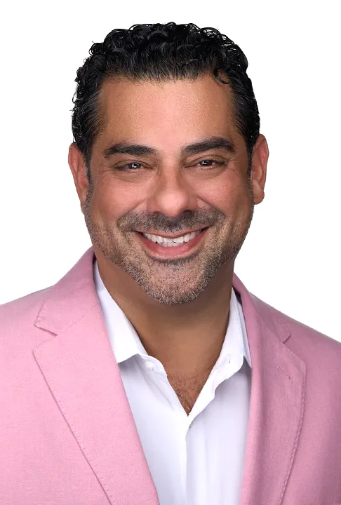 A man with black hair wears a pink blazer as he smiles for a professional headshot while standing in front of a white backdrop in Raleigh Nc