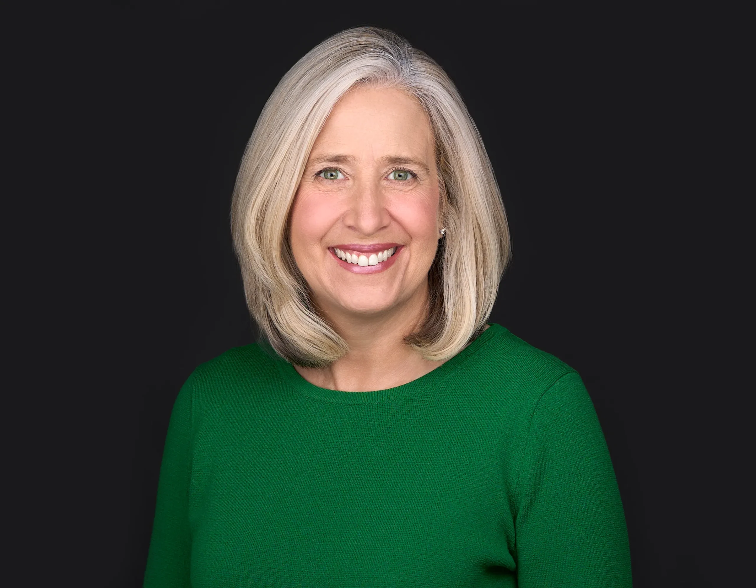 A women with gray hair and a bright green sweater smiles beautifully while posing for a professional headshot in front of a black background in Raleigh Nc