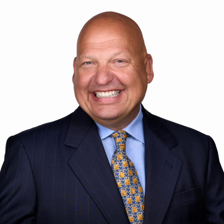 A bald man in a dark pinstripe suit, blue shirt, and patterned tie smiles at the camera against a plain white background.