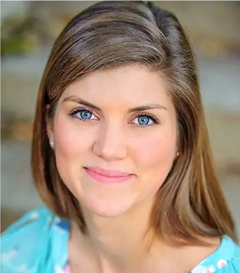 A woman with straight, light brown hair and blue eyes smiles softly at the camera. Captured by a skilled headshots photographer, she wears a light blue top and is photographed against a blurred background.
