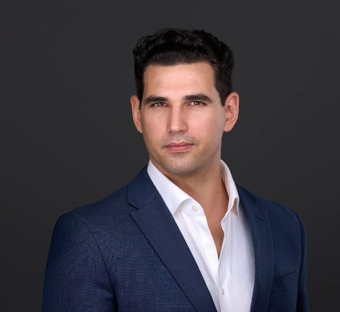 Man in a navy suit and white shirt poses against a plain dark background, looking directly at the camera with a neutral expression.