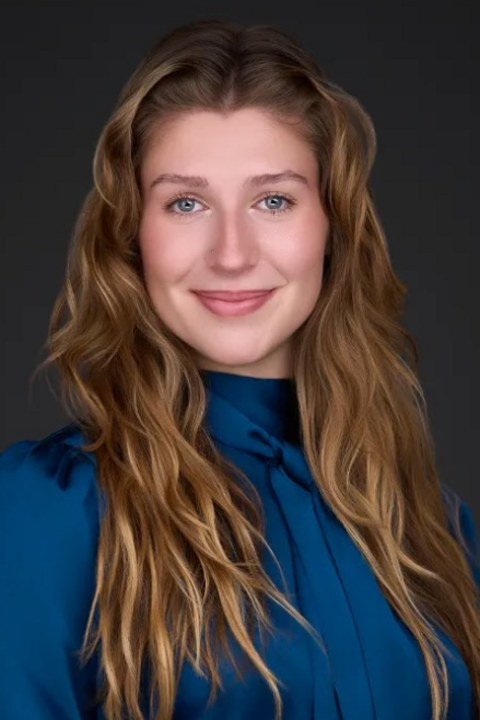 Woman with long wavy light brown hair, wearing a blue blouse, smiling slightly, posing in front of a plain dark background.