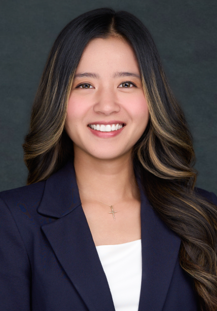 A woman with long, wavy dark hair wears a navy blazer over a white top and a cross necklace, smiling in front of a dark grey background.