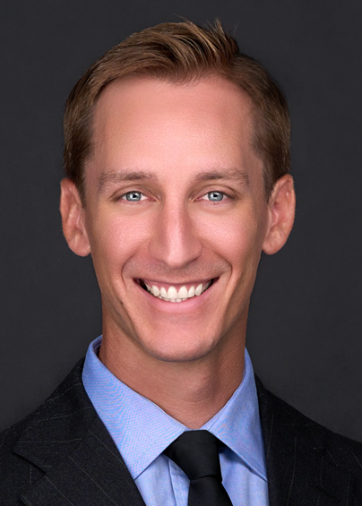 A man in a suit and tie smiles at the camera against a plain dark background.
