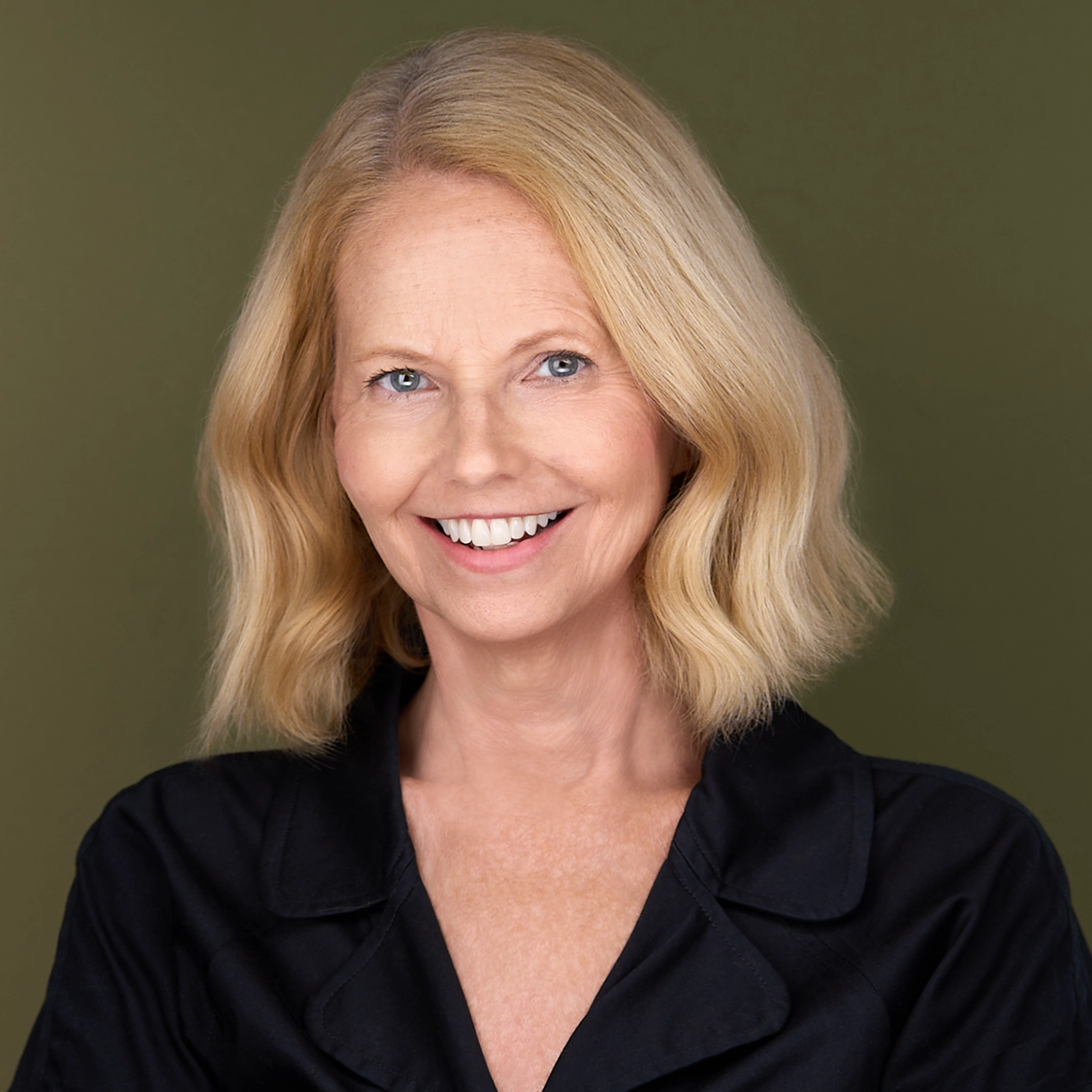 Smiling middle-aged woman with blonde shoulder-length hair wearing a black collared shirt, posed against a plain olive green background.