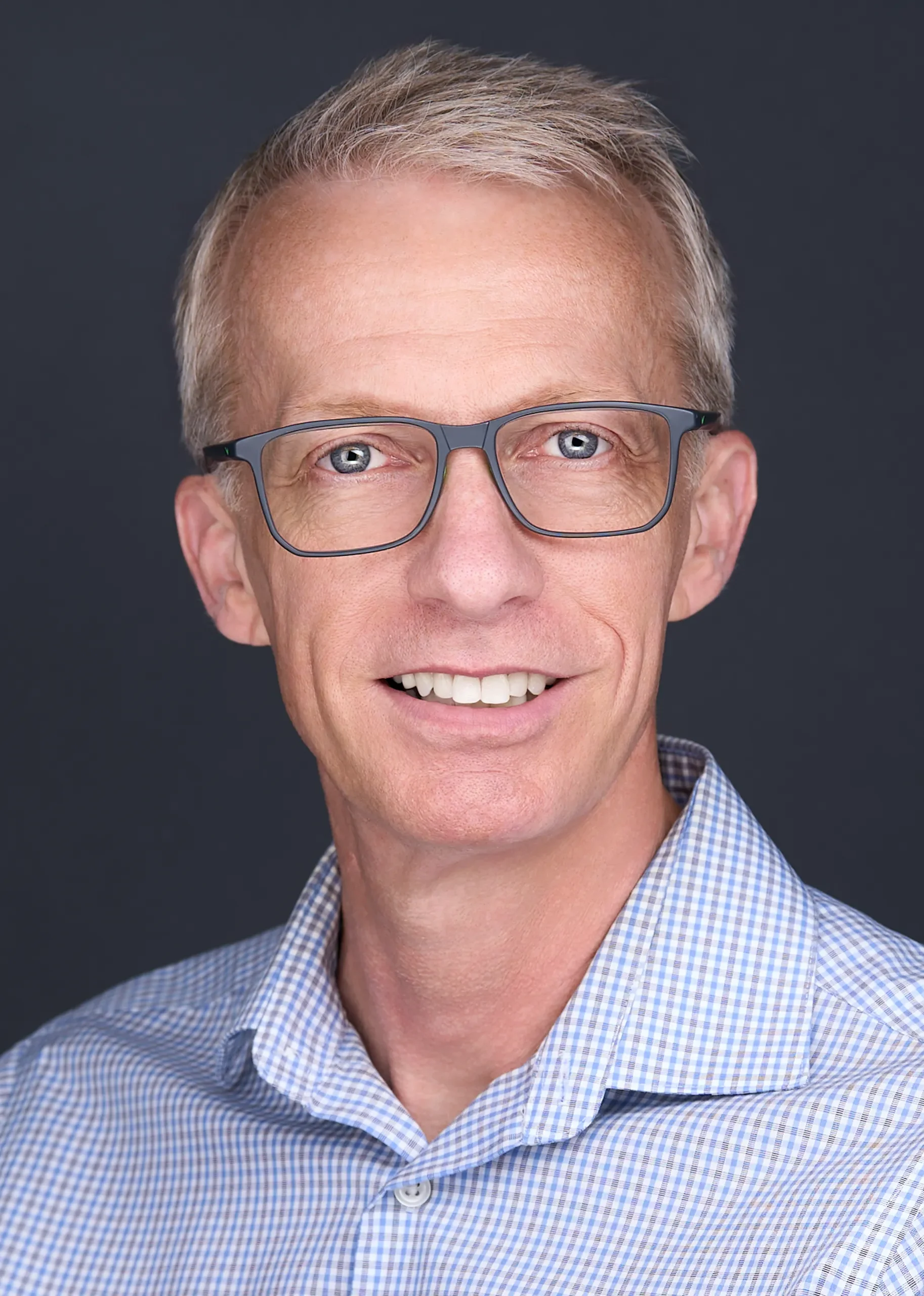 A man with blue framed glasses and gray hair sits for a professional headshot while standing in front of a grey backdrop in Cary Nc