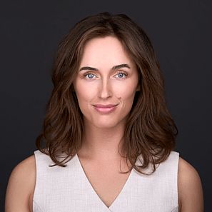 A young woman with brown hair wearing a beige sleeveless vest sits for a professional portrait .