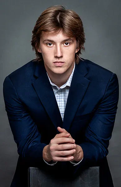 A young man dressed in a navy blue jacket and light blue shirt leans on a posing box as he stares into the camera for a personal branding headshot in Cary Nc