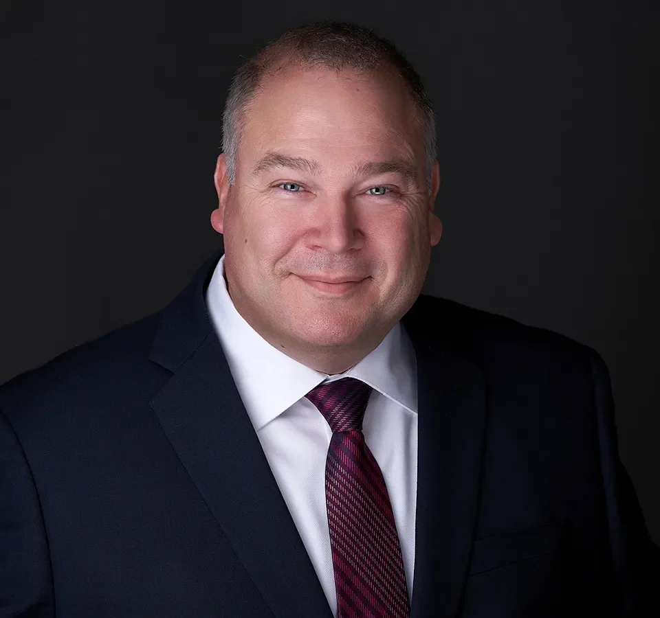 A man wearing a black suit and red tie gently smiles while posing in front of the camera in the studio of Sarah Anne Wilson in Cary Nc