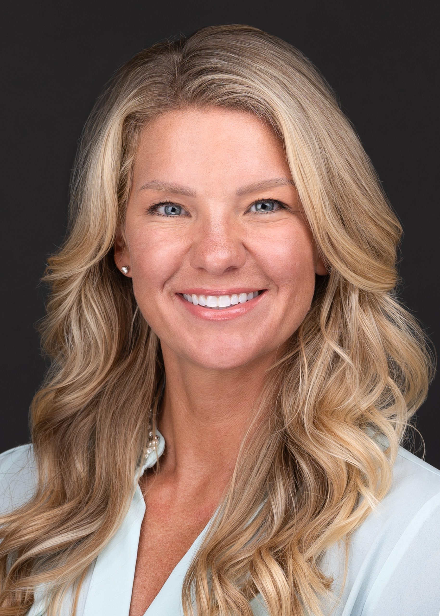 A woman with long blonde hair wearing a light blue blouse poses for her corporate headshot.