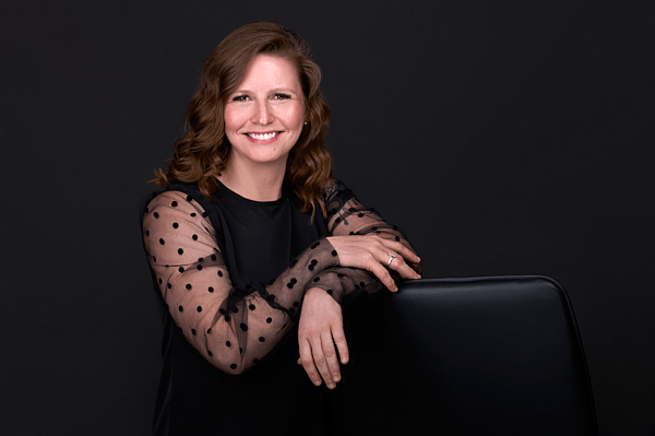 A woman with wavy brown hair smiles while leaning on the back of a chair. She is wearing a black top with sheer, polka-dotted sleeves. The background is black, highlighting the elegance perfect for branding photography.