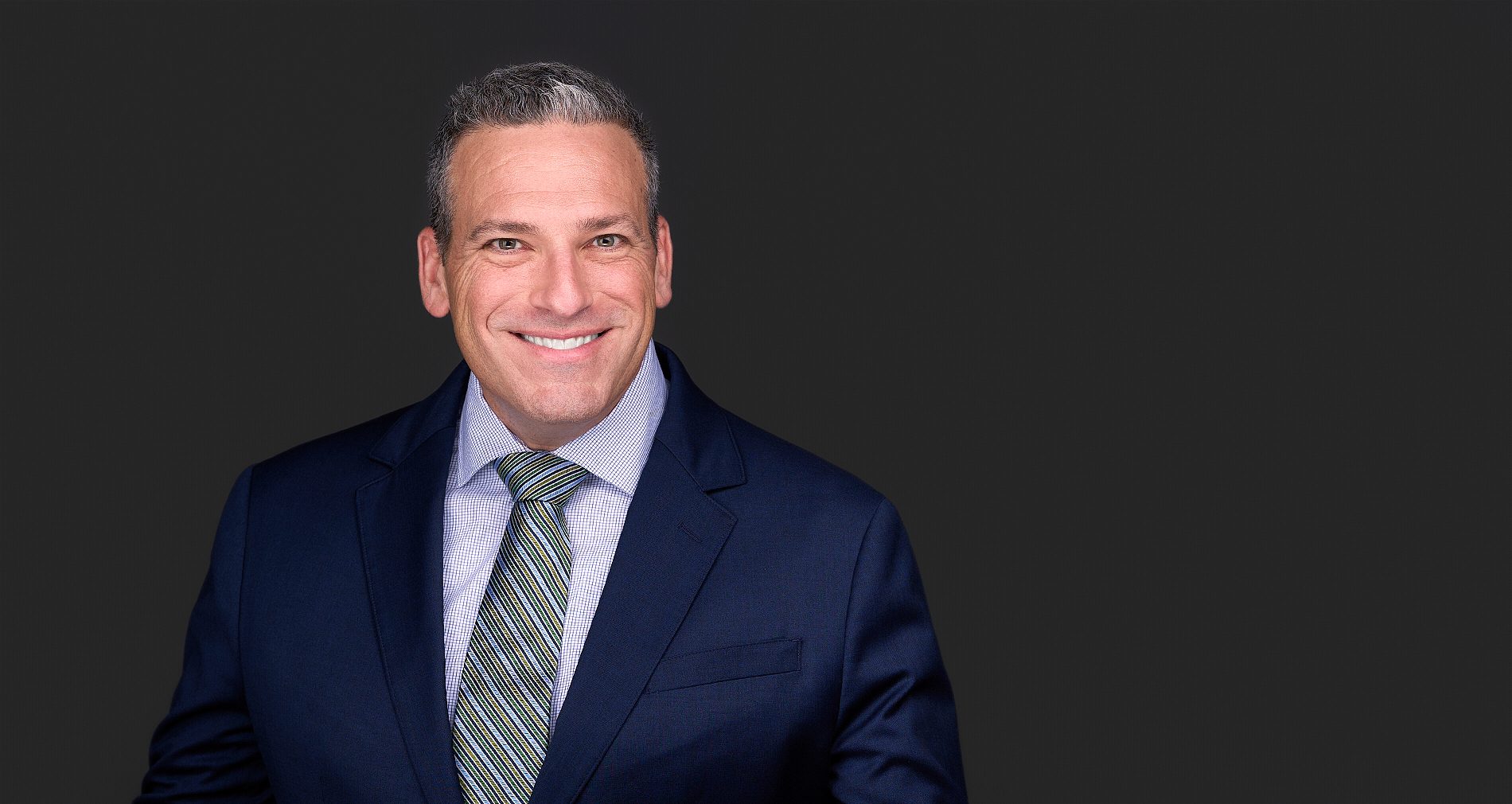 A man wearing blue suit and striped tie smiles as he poses for a lawyers headshot in Cary NC