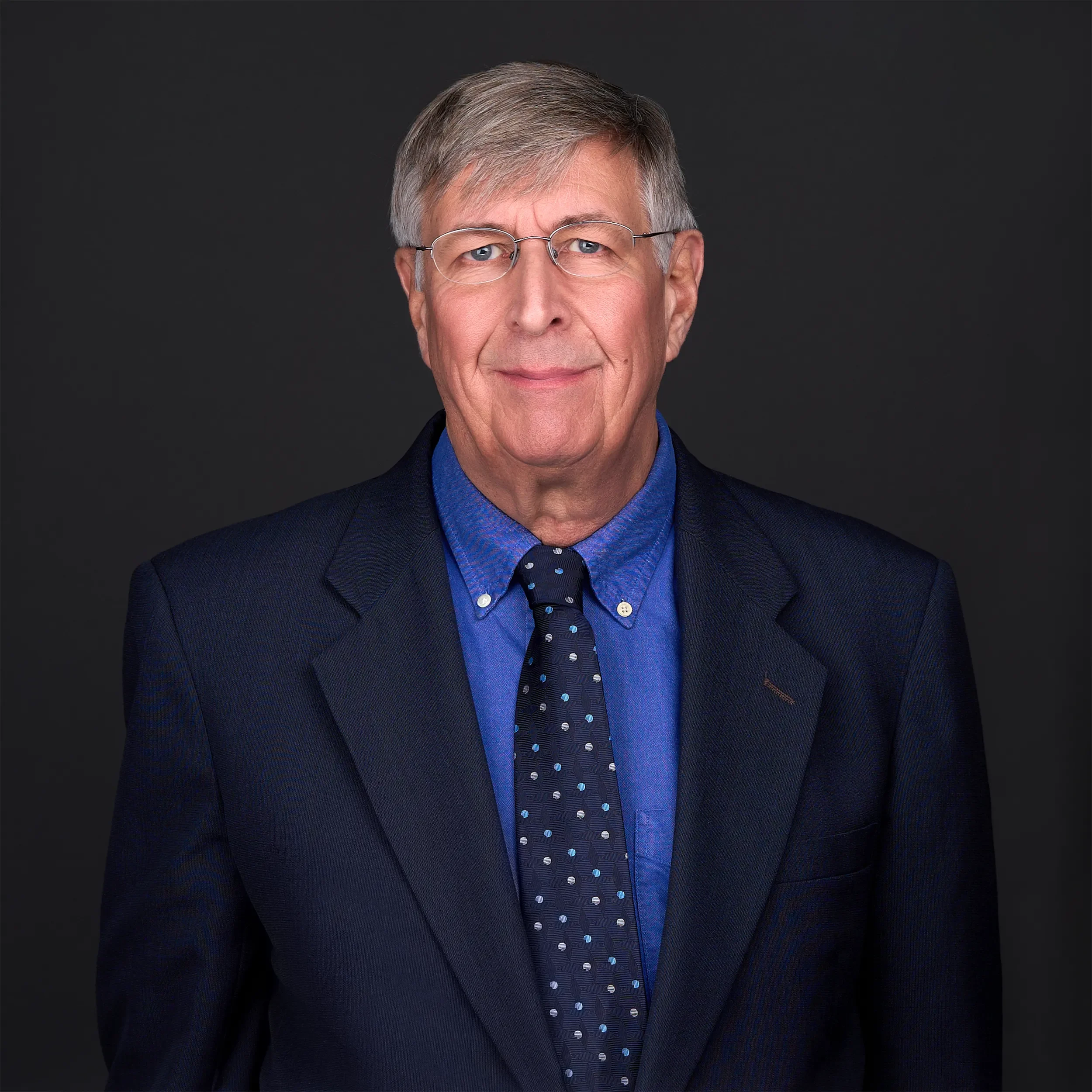 An elderly man with glasses wears a bright blue shirt and navy jacket as he stands in front of a camera in a photography studio in Cary Nc