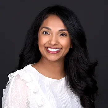 A woman with long, dark hair and a white blouse smiles confidently in her professional headshot against a dark background.