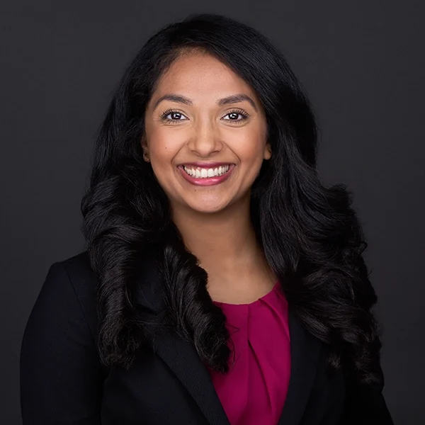 womens-professional-headshot Young women posing in front of a black backdrop with long black hair and wearing a red bouse and black jacket smiles warmly for her headshots in Cary NC