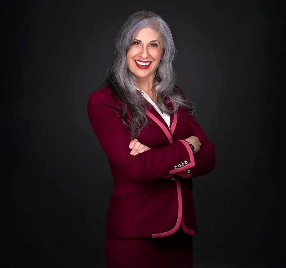 A woman with long gray hair wearing a maroon blazer stands with arms crossed, smiling in her professional headshot against a dark background.