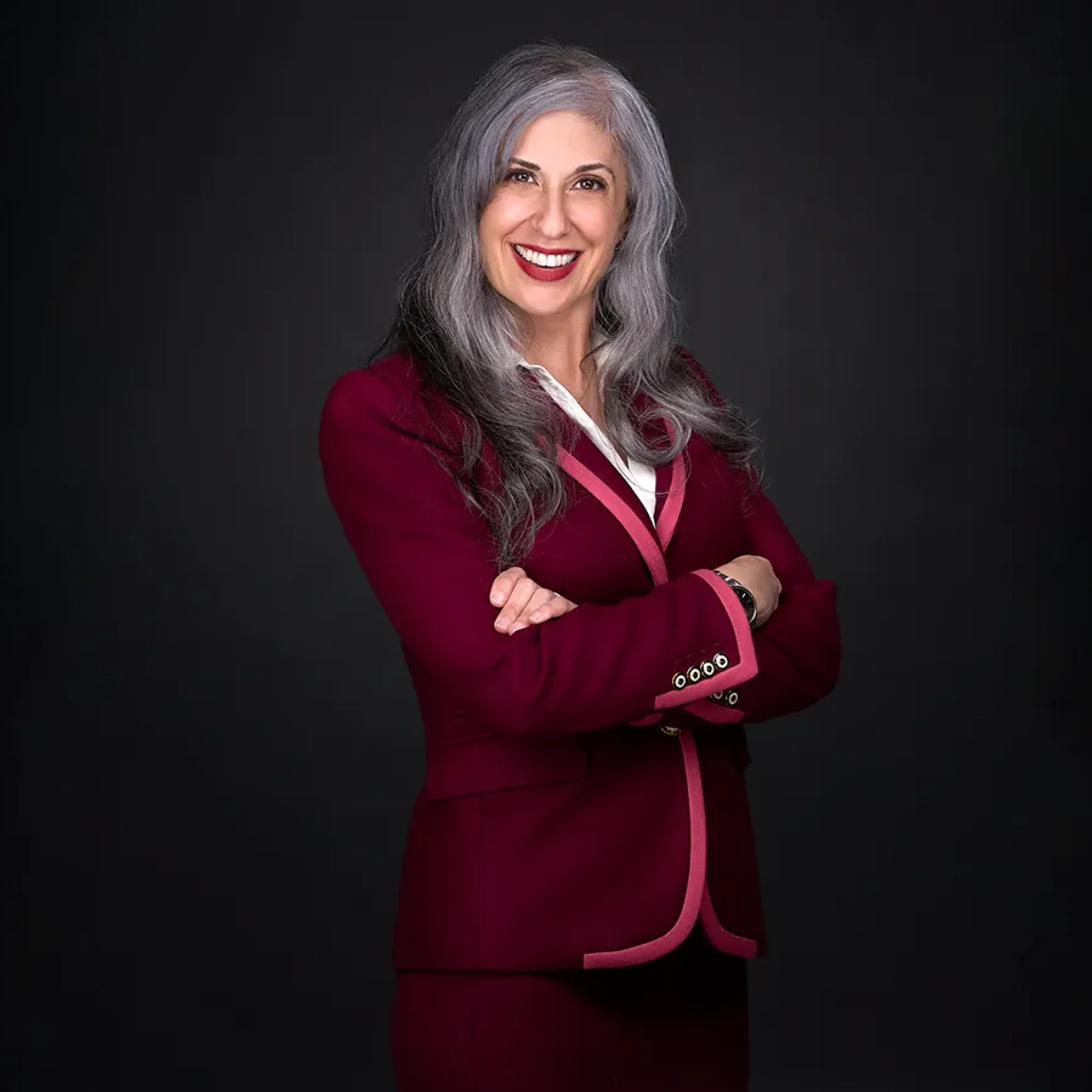 A woman with long gray hair wearing a maroon blazer stands with arms crossed, smiling in her professional headshot against a dark background.