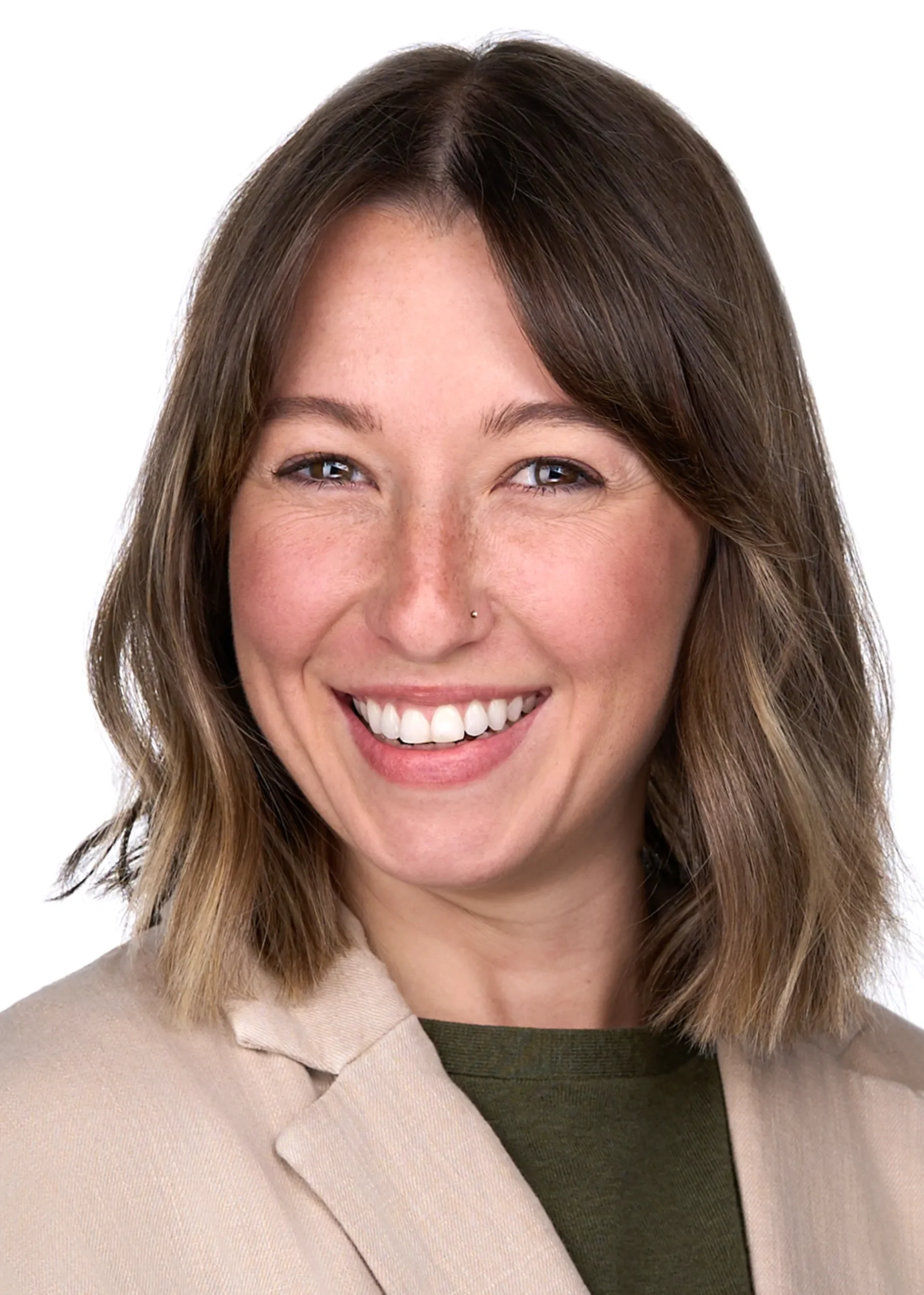 A young woman with shoulder length brown hair wearing a forest green sweater and tan blazer smiles for the camera as she stands in front of a white backdrop in the studio of Sarah Anne Wilson Photography