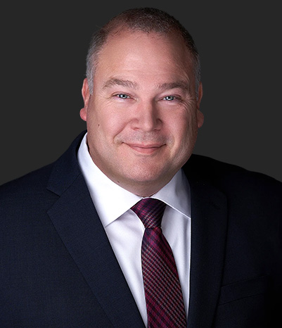 A middle-aged man wearing a dark suit, white shirt, and plaid tie, smiling at the camera against a plain dark background.